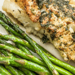 baked cod with garlic herb butter on top next to asparagus spears on white plate