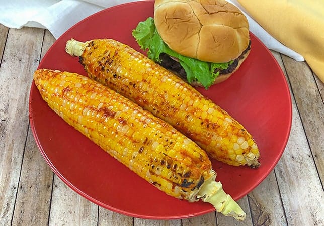 two ears of grilled corn on red plate with a hamburger on the side