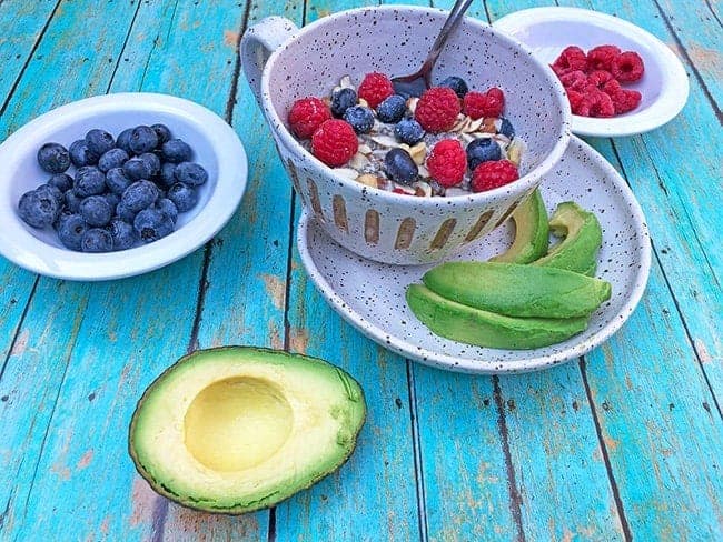 low carb oatmeal in white bowl with blueberries and raspberries on top and avocado slices on side