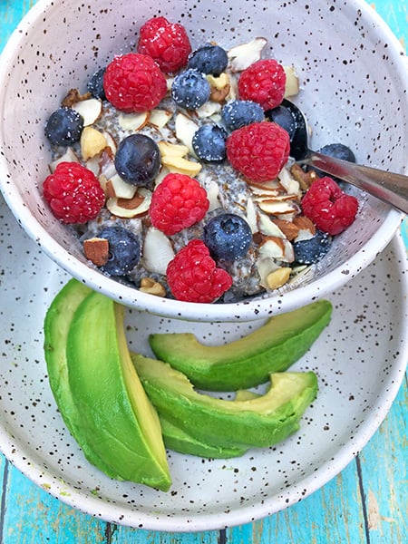 keto low carb oatmeal in a white bowl topped with blueberries, raspberries, and almond slices. Sliced avocado on the side.