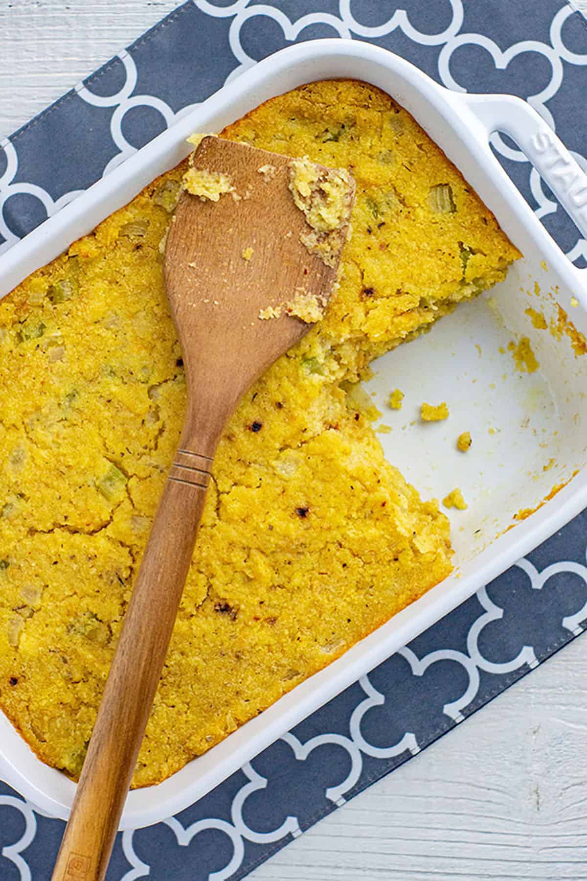 baked cornbread dressing in white rectangular casserole dish with wooden serving spatula on top. One serving of dressing from the corner is missing from the casserole dish.