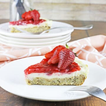 slice of cheesecake with strawberry on top on white plate with fork. Pink napkin on the side. Another slice of cheesecake in the background.