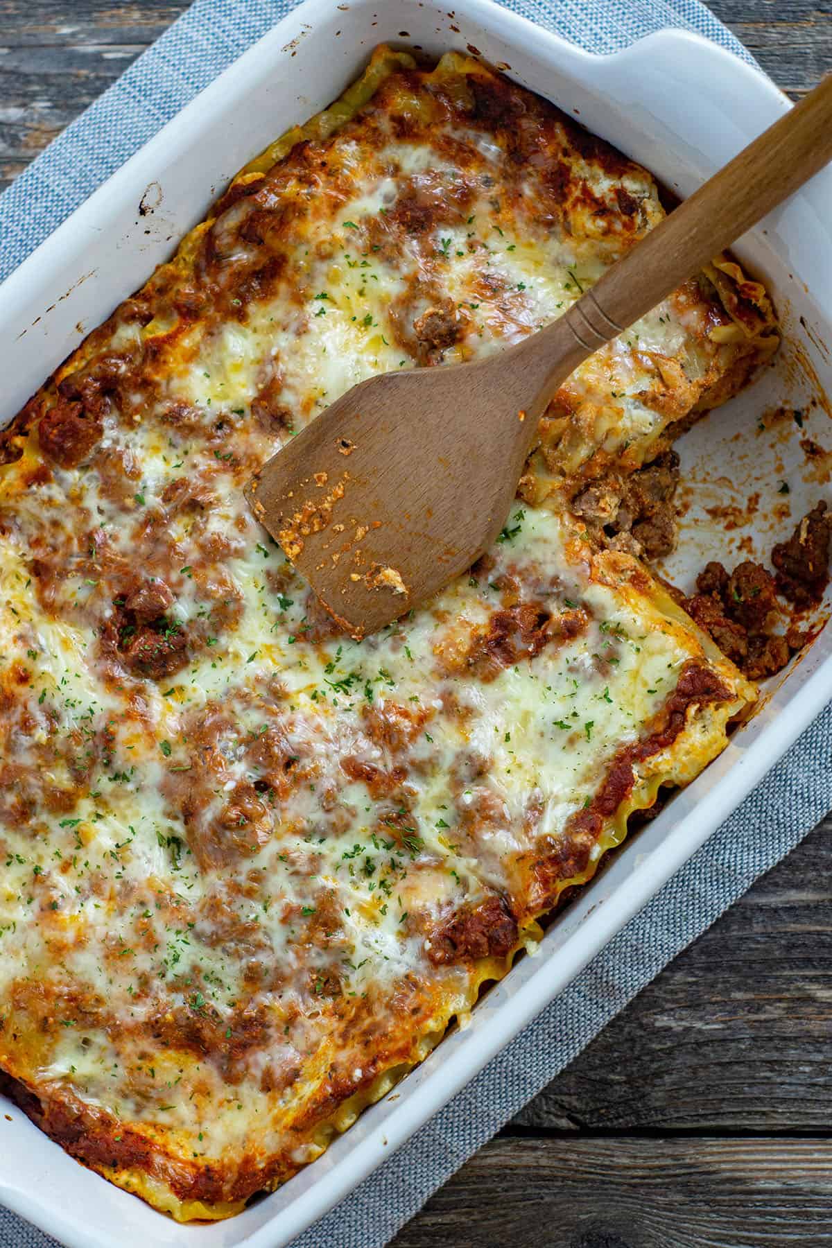 baked lasagna in white rectangular dish with slice removed wooden spoon on top