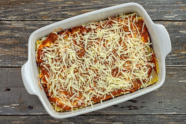 3 layer assembled meat lasagna in a white casserole dish with cheese on top prior to baking 