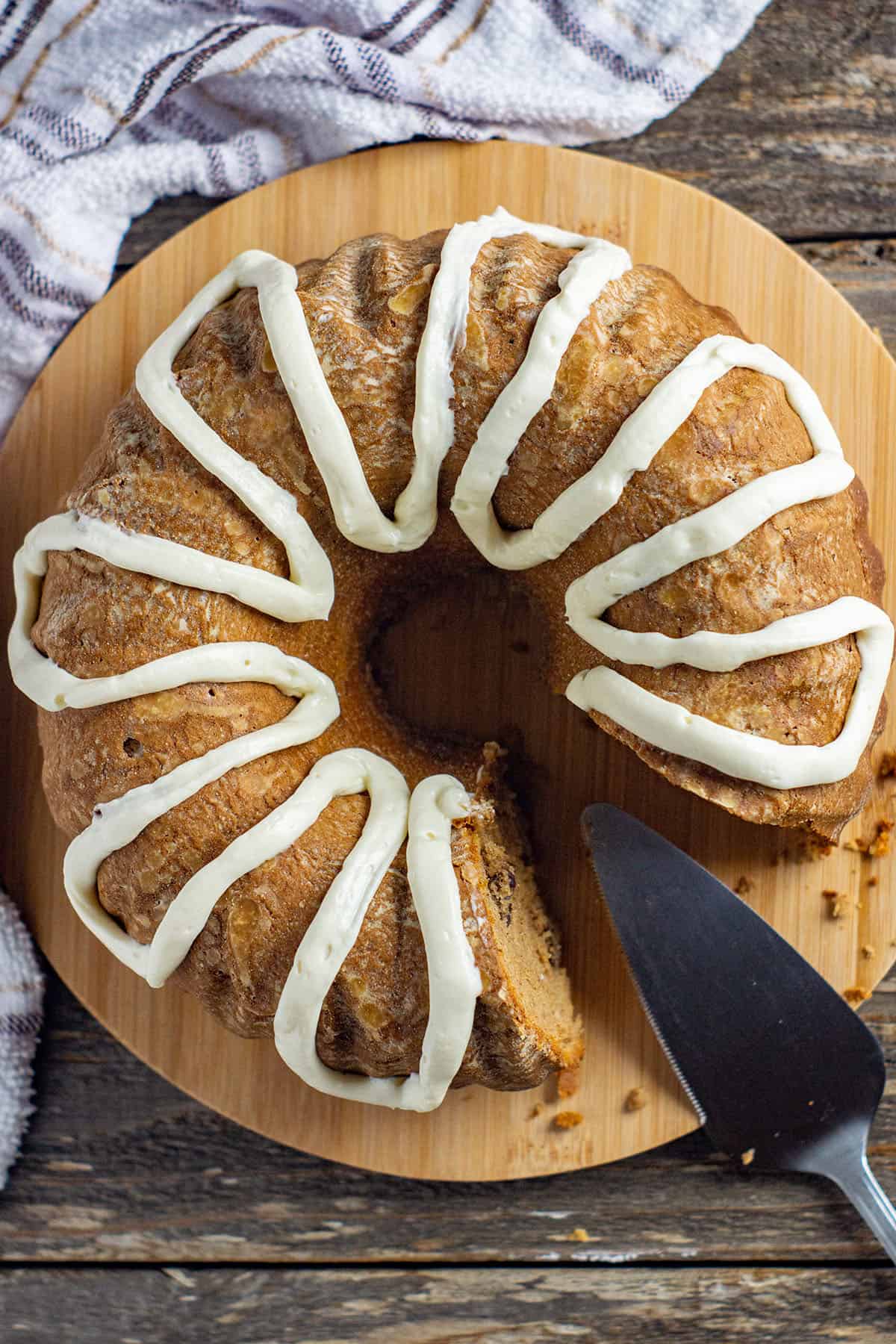 overhead view of baked and frosted cake on round wooden cake platter with one piece of cake removed.  silver cake spatula resting on platter.