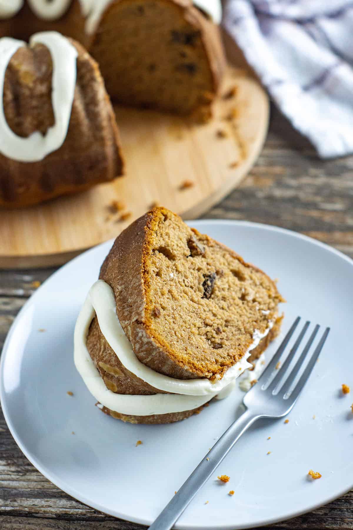 slice of pumpkin spice bundt cake on a white dessert plate with silver fork and remaining bundt cake in the background
