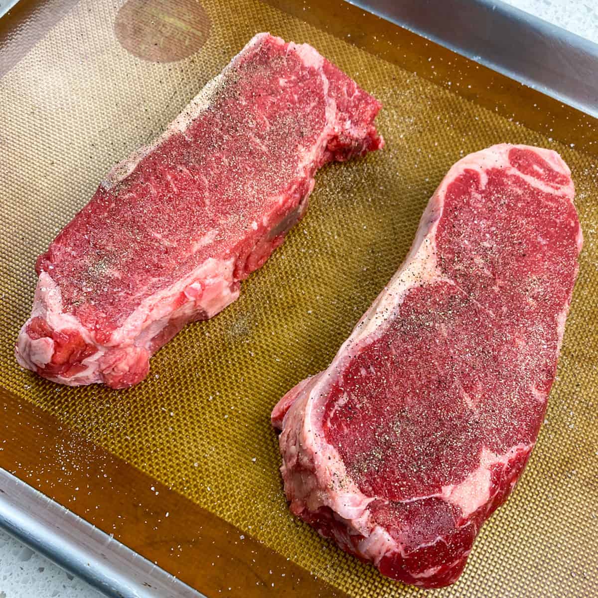 two uncooked seasoned new york strip steaks on baking sheet