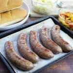 cooked bratwurst sausages in square pan with buns, sauerkraut, and bell pepper onion mixture on the side
