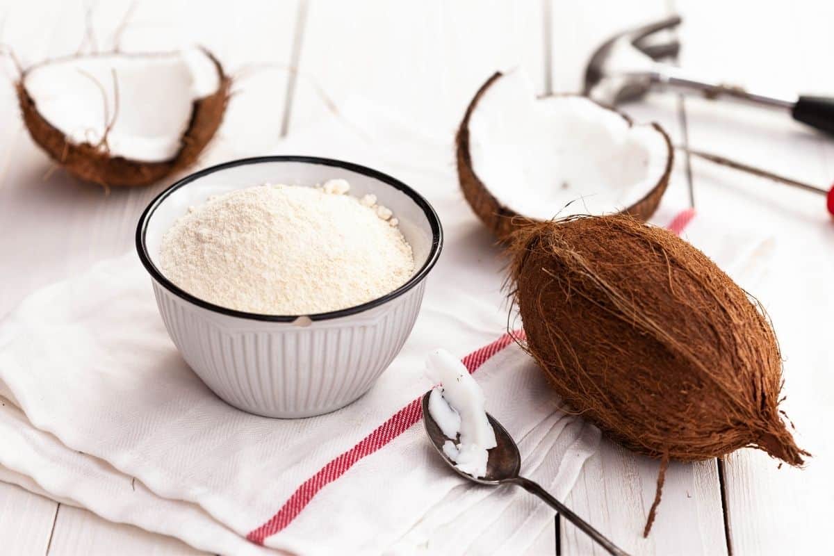 coconut flour in white bowl with whole coconut next to it and two coconut halves with a hammer in the background