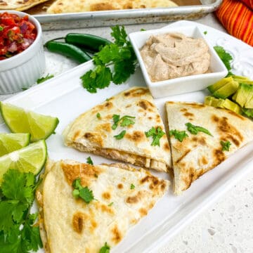 cooked quesadillas cut into thirds on white rectangular plate with lime wedges and seasoned sour cream