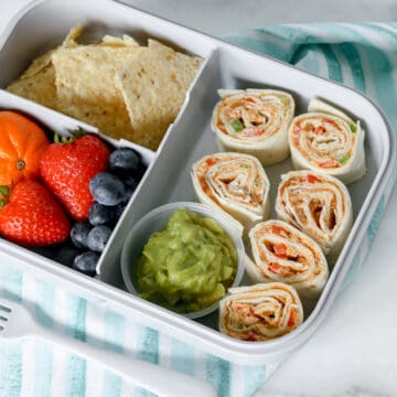 sliced taco pinwheels in lunch box with guacamole, chips, strawberries, and blueberries