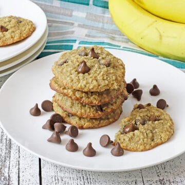 baked cookies stacked on a white plate with chocolate chips scattered around and fresh bananas in the background