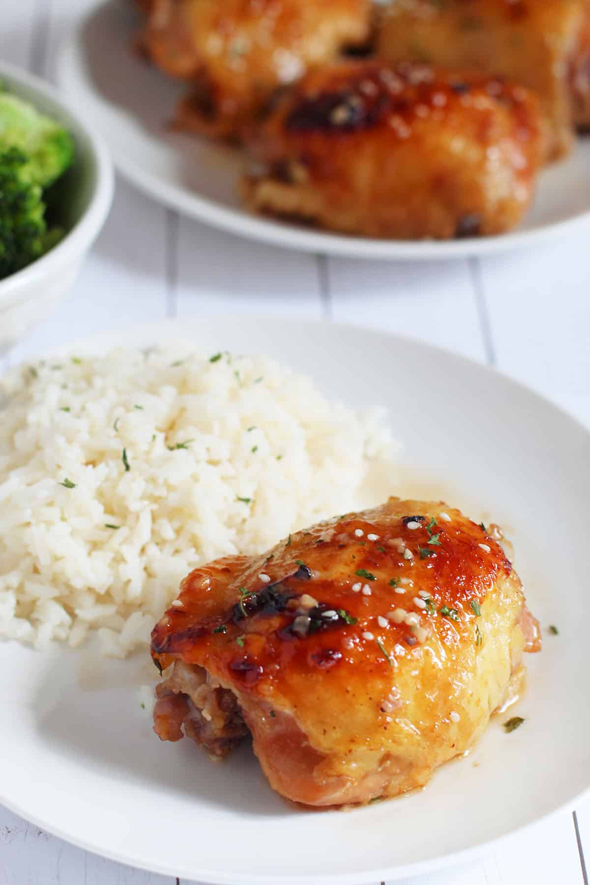 Baked chicken thigh and white steamed rice on white place with broccoli in background.