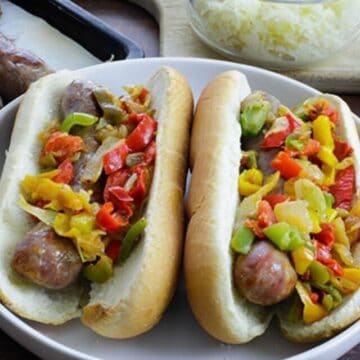 Two baked brats in buns topped with sauteed bell peppers and onions.