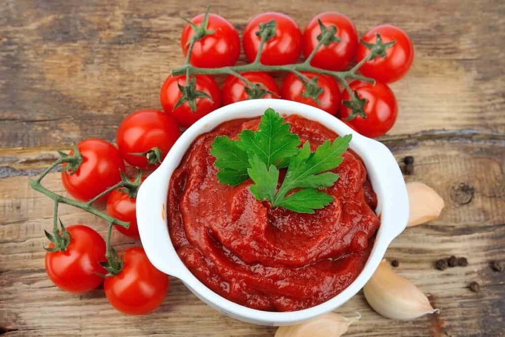 tomato paste in white bowl with parsley sprig on top and tomatoes on the vine next to the bowl on top of wooden counter top