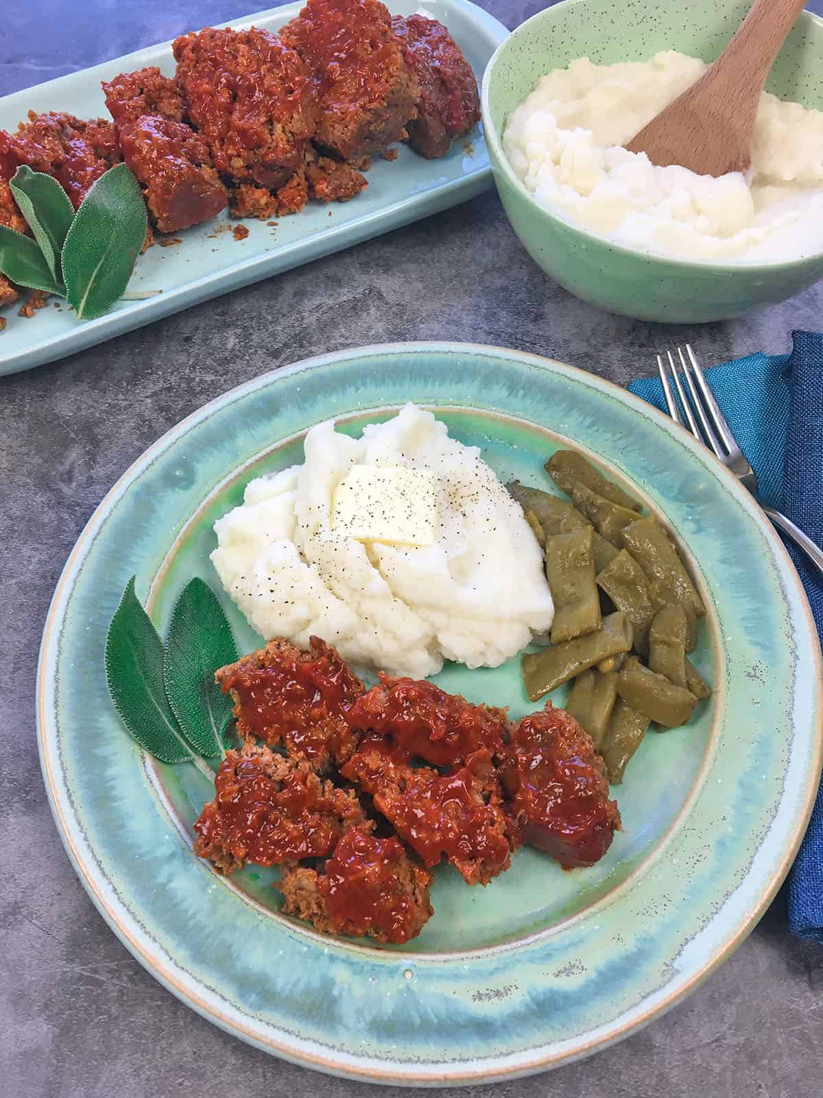 Sliced meatloaf on teal blue plate with a serving of mashed potatoes and green beans on the side.