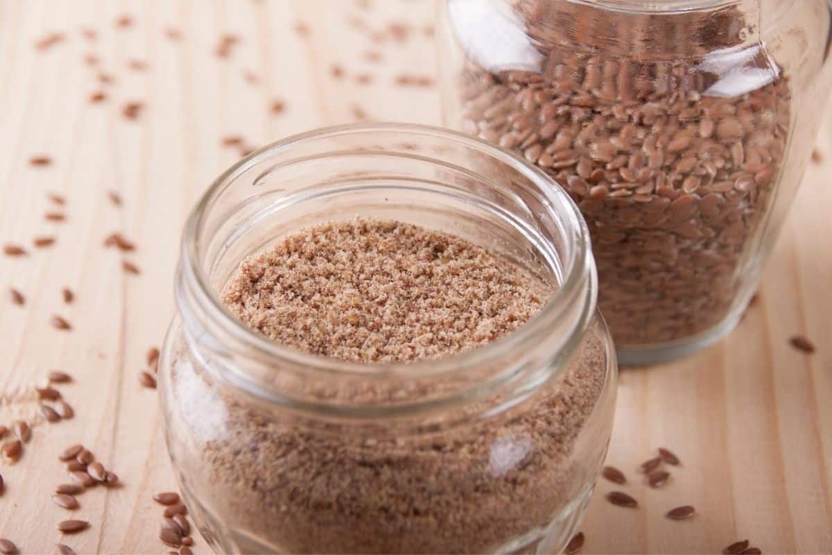 ground flax meal in mason jar with whole flax seeds in another mason jar in background.  Jars on wooden counter top with whole flax seeds scattered around.