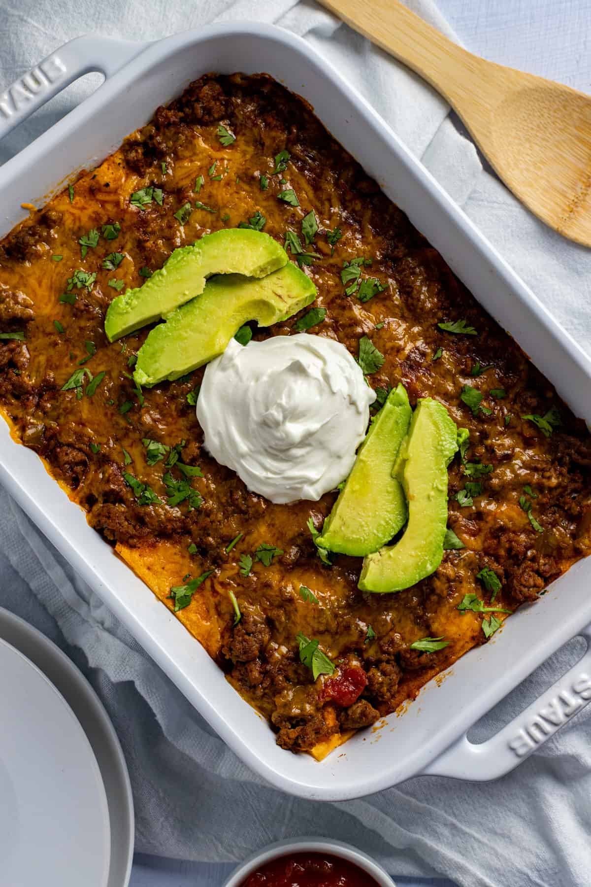 baked casserole in white rectangular dish.  Topped with sliced avocados and dollop of sour cream.  Wooden spoon on the side.