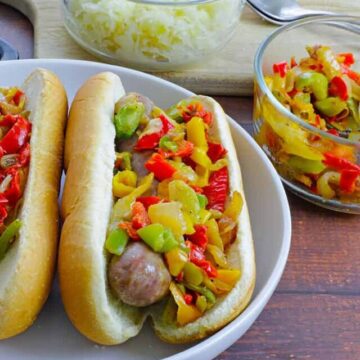 brats in buns topped with red, green, and yellow bell pepper strips on top. Sauerkraut in bowl in background.