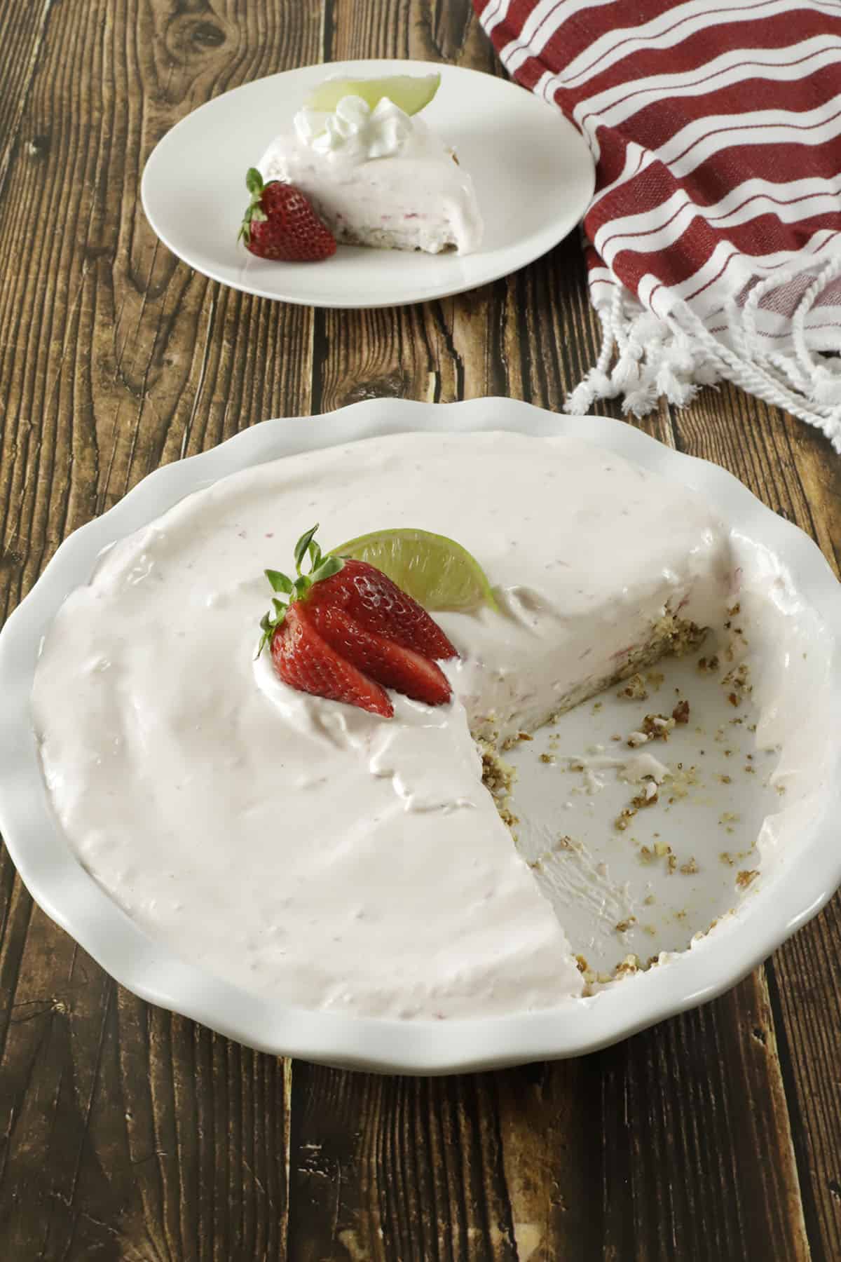 Whole pie with slice removed, exposing pretzel crust, in a white fluted pie dish garnished with lime wedges and strawberry slices.