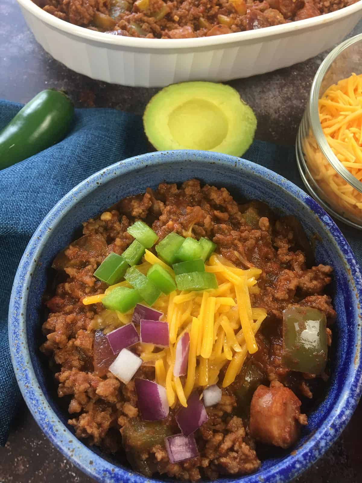 Cooked chili topped with shredded cheddar cheese, diced onions and jalapenos with avocado half and whole jalapeno in background.