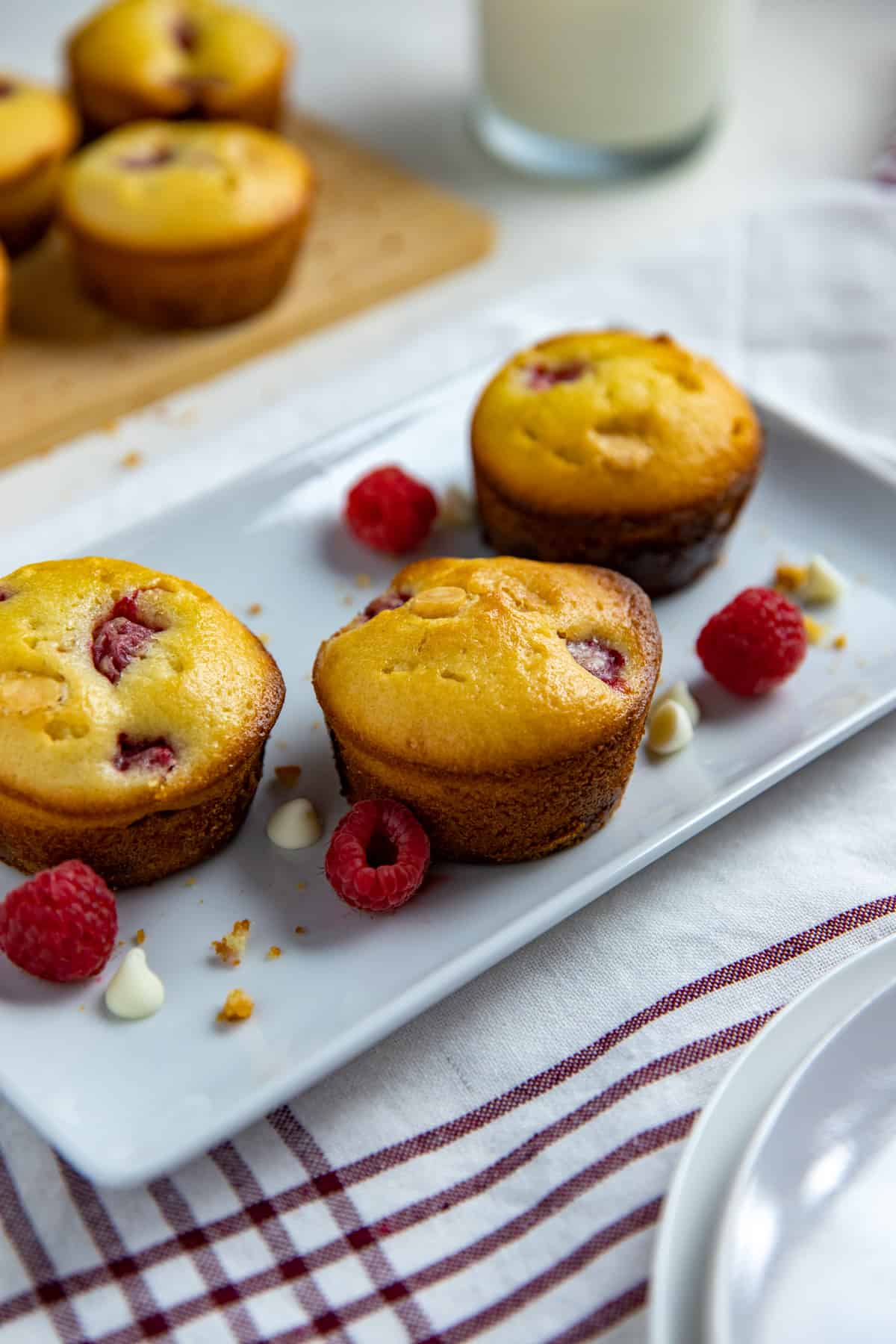 three baked muffins on white rectangular dish with raspberries and white chocolate chips scattered around the muffins