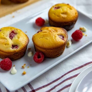 three baked muffins on white rectangular dish with raspberries and white chocolate chips scattered around the muffins