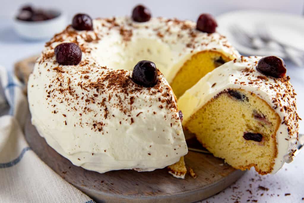 A bundt cake with white frosting, chocolate shavings, and cherries on top. One slice is cut and slightly removed, revealing a moist yellow interior with embedded cherries. The cake is on a wooden board with a checkered cloth partially visible.
