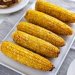 Five ears of grilled corn on white square platter with hamburger in background
