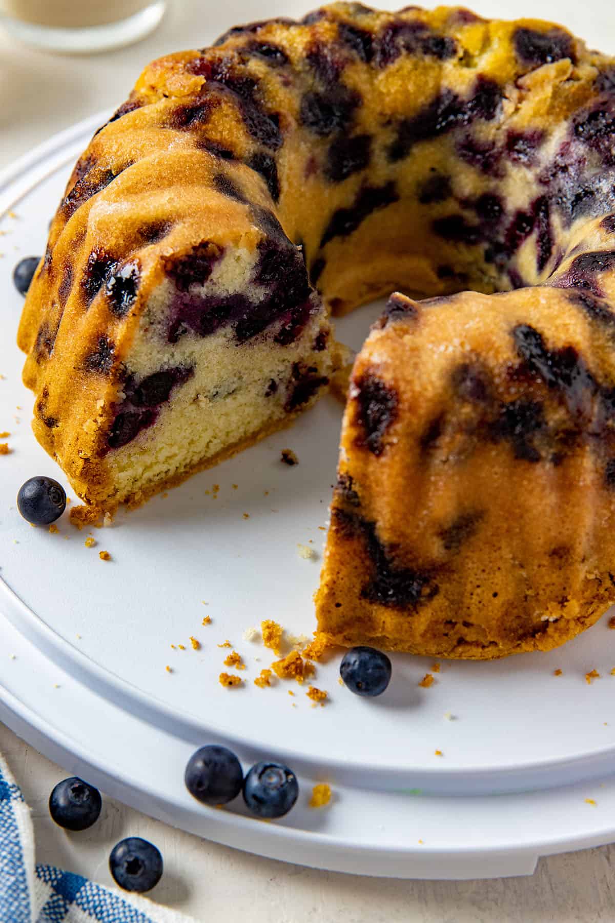 baked cake with one slice removed.  Cake on white cake holder with fresh blueberries scattered around.