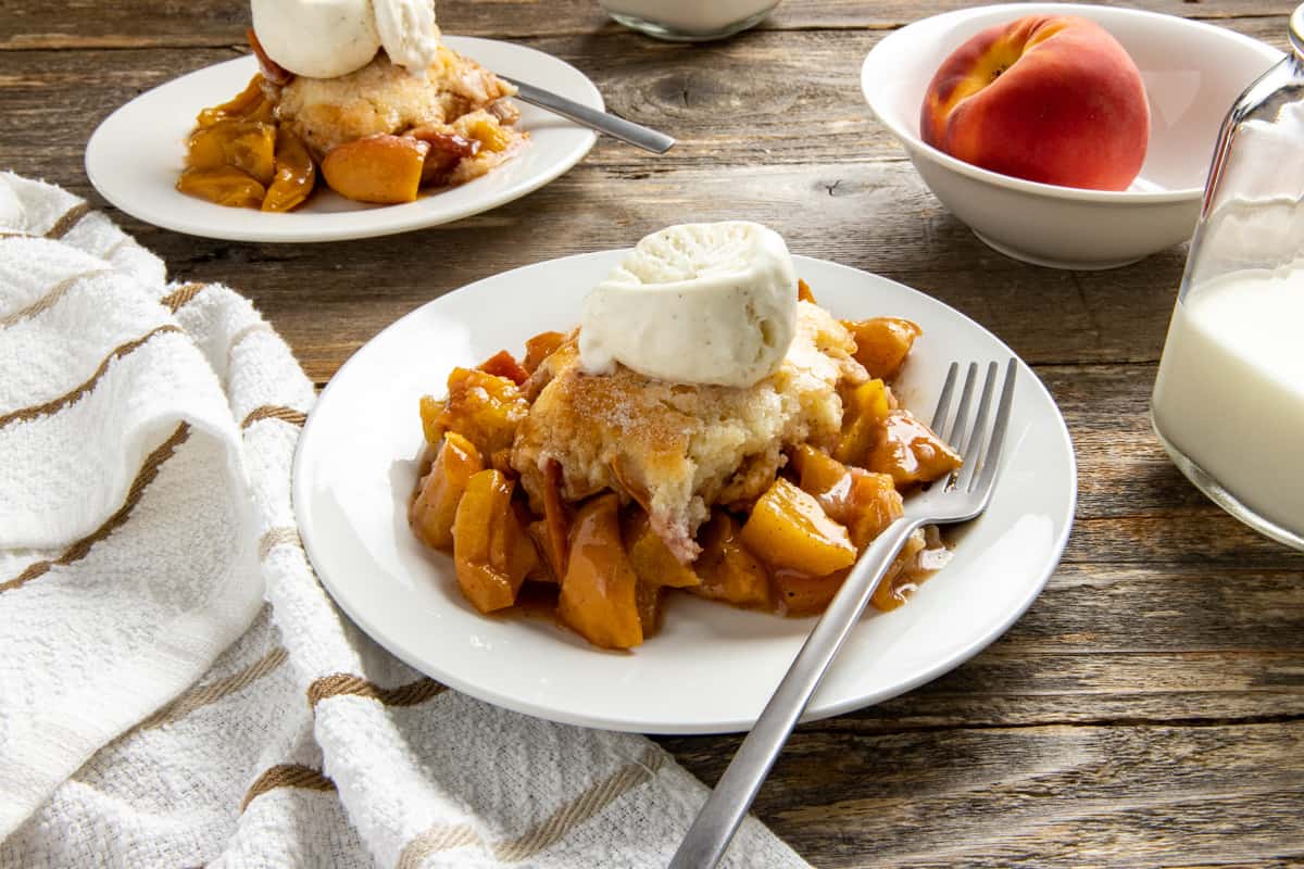 peach cobbler and ice cream in a white bowl with fork.  another serving in the background.  fresh peach on the side.