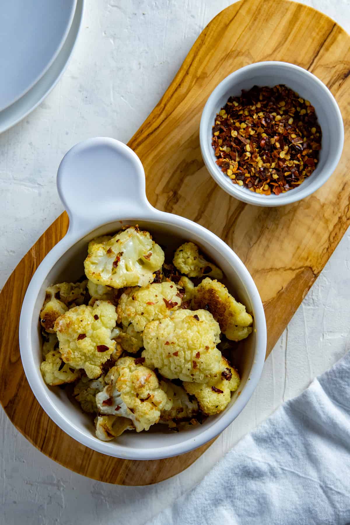Bowl of roasted parmesan cauliflower with a small bowl of red pepper flakes on the side.  Both bowls sitting on a wooden platter.