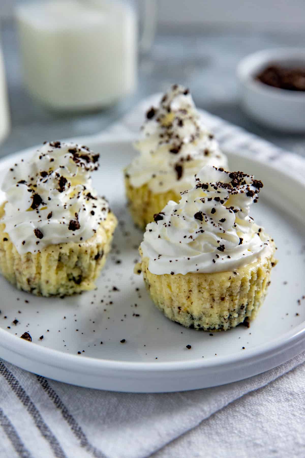 Three mini cheesecakes with whipped cream topping and Oreo crumbs sprinked on top.  Cheesecakes on white plate with cup of milk in background and a small bowl of crushed Oreo cookies.