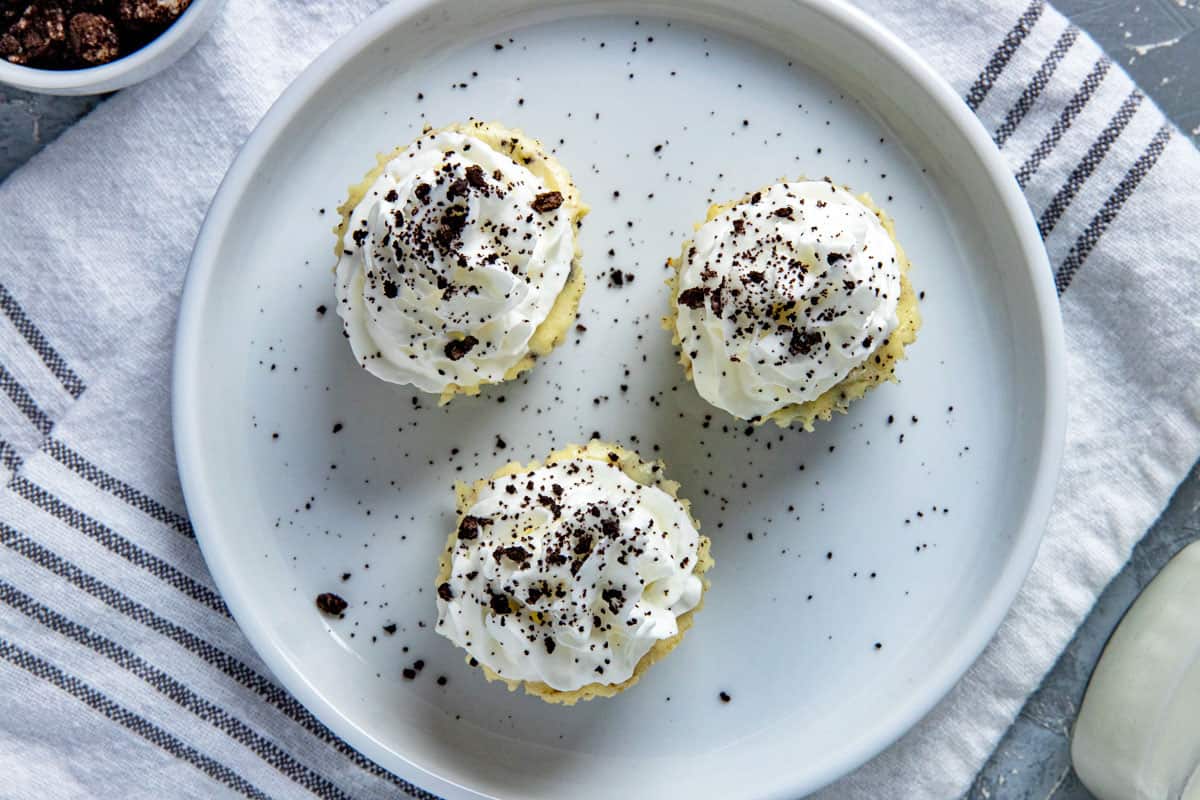 Three mini cheesecakes with whipped cream topping and Oreo crumbs sprinked on top. Cheesecakes on white plate with cup of milk in background and a small bowl of crushed Oreo cookies.