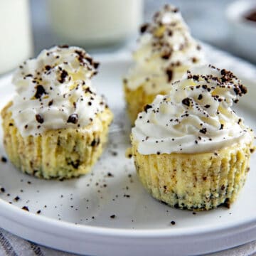 Three mini cheesecakes with whipped cream topping and Oreo crumbs sprinked on top. Cheesecakes on white plate with cup of milk in background and a small bowl of crushed Oreo cookies.