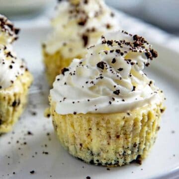 Three mini cheesecakes with whipped cream topping and Oreo crumbs sprinked on top. Cheesecakes on white plate with cup of milk in background and a small bowl of crushed Oreo cookies.