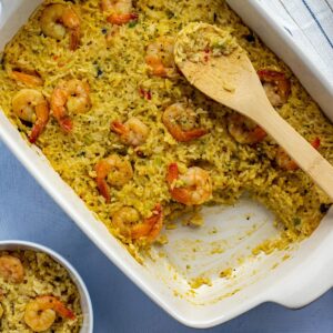 Baked Shrimp and Rice Casserole in 9-inch by 13-inch white casserole dish with a serving removed and placed in a small white bowl. Wooden spoon on the side.