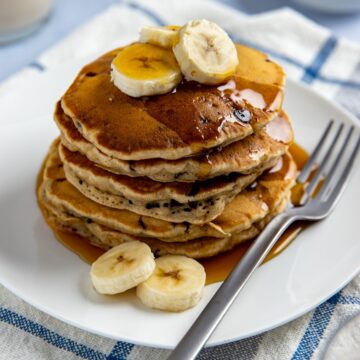 Five chocolate chip banana pancakes stacked on white plate with slices of fresh bananas on top and sides. Drizzled with syrup. Fork on plate.