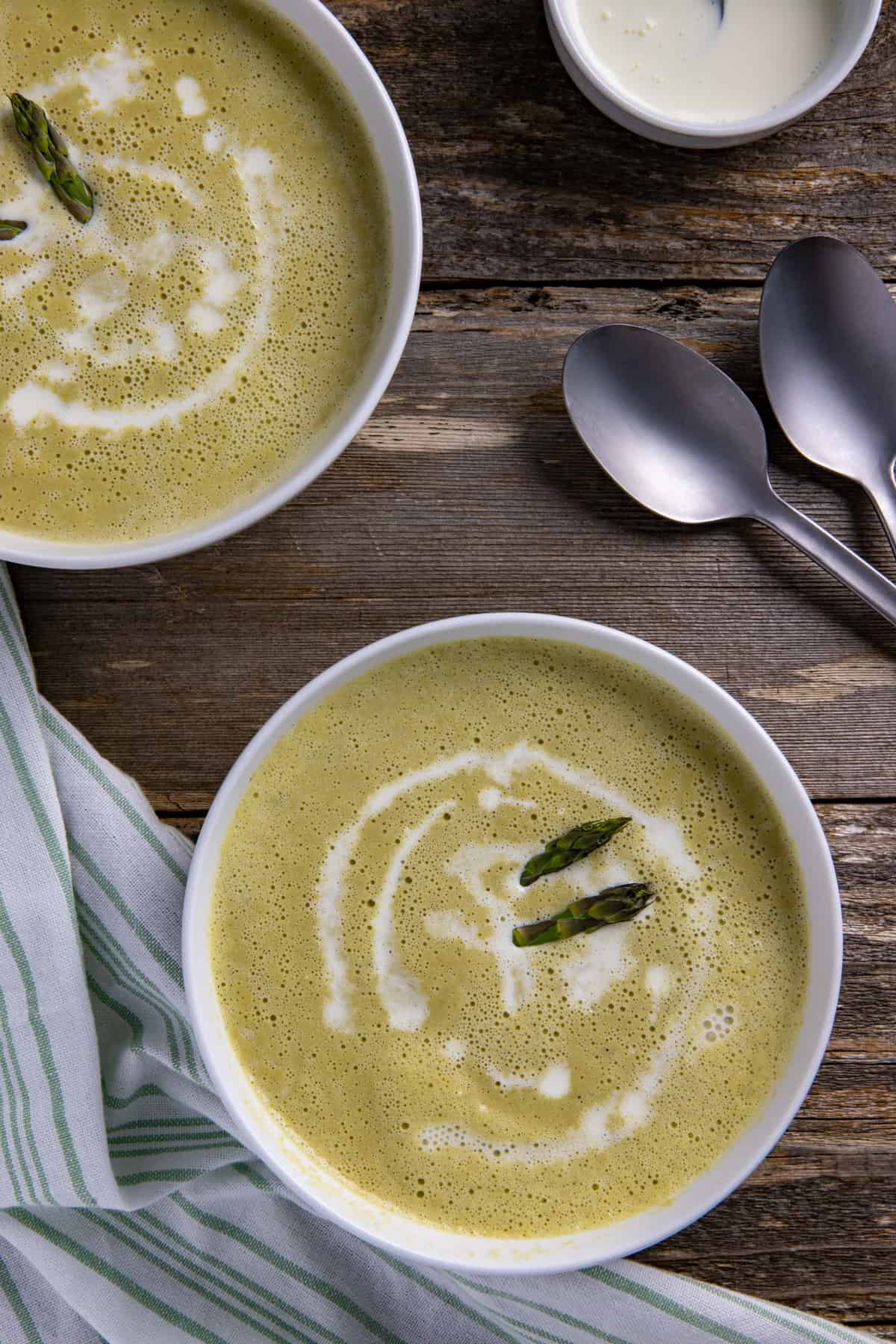 Two white bowls filled with soup garnished with two fresh asparagus tips and a swirl of heavy cream.