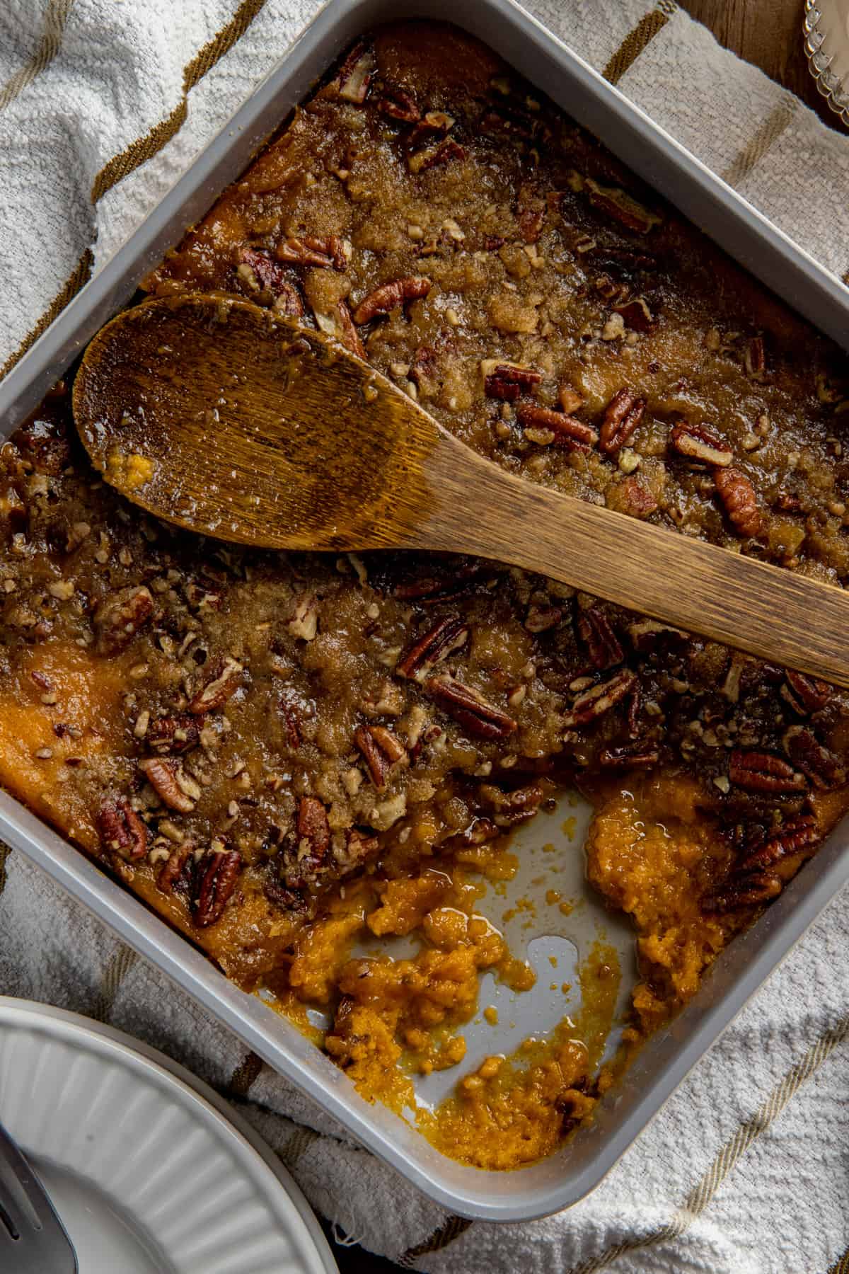 Baked sweet potato casserole in square aluminum pan. Wooden spoon on top of casserole with one serving removed.