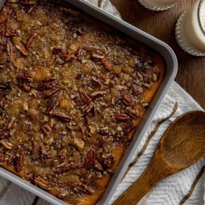 Baked sweet potato casserole in square aluminum pan. Wooden spoon on white napkin next to casserole dish.