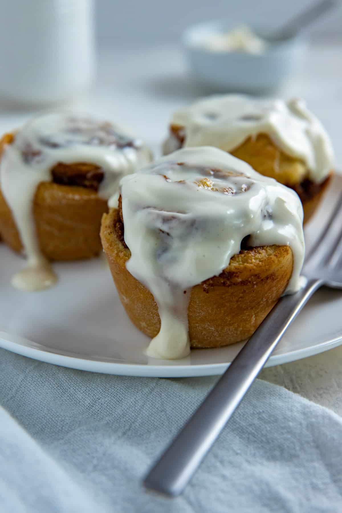 Three baked cinnamon rolls with cream cheese frosting on white round plate with fork.