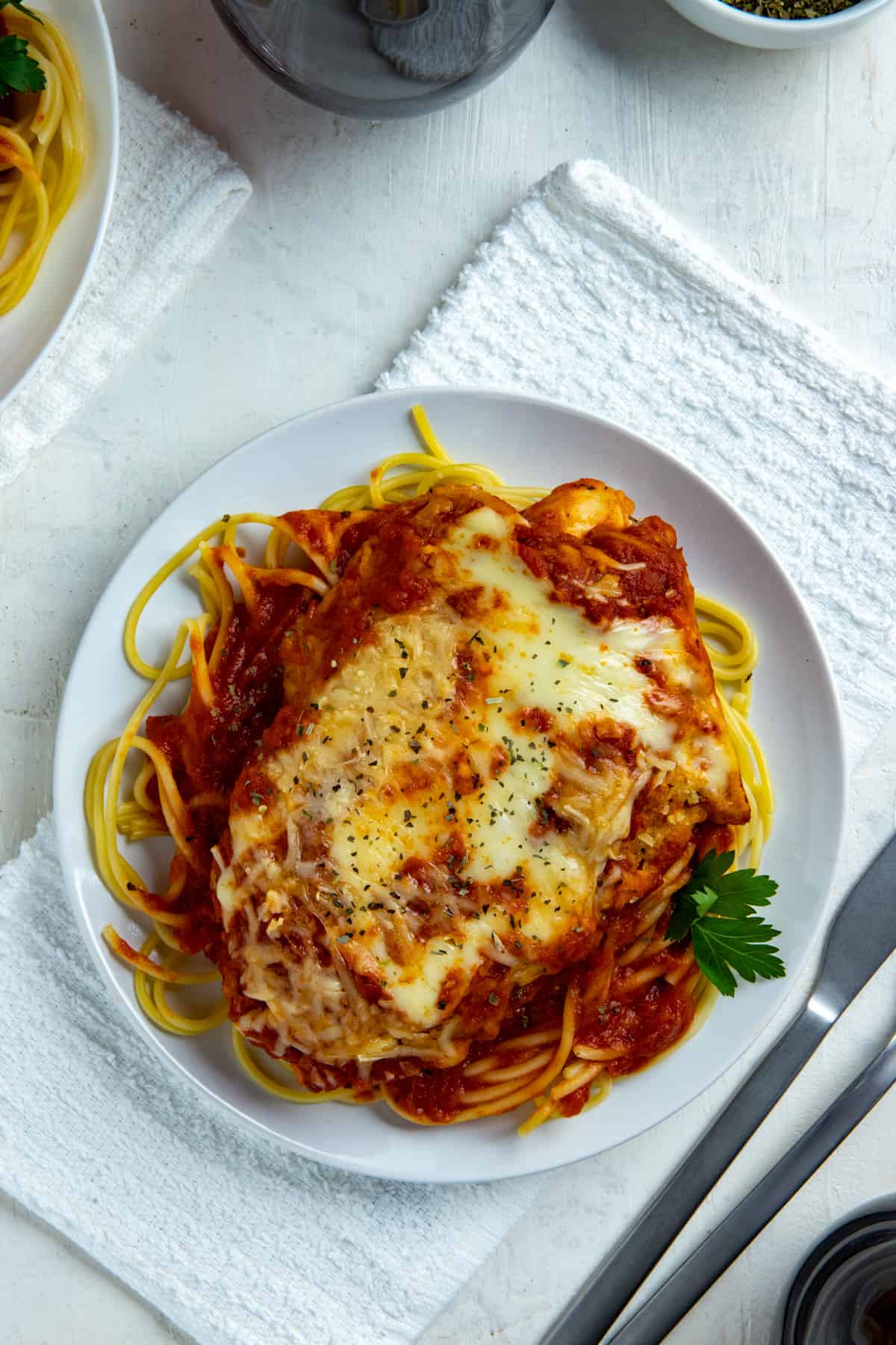 Chicken Parmesan over Spaghetti on round white plate with fork and knife on the side.