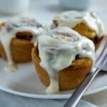 Three baked cinnamon rolls with cream cheese frosting on white round plate with fork.