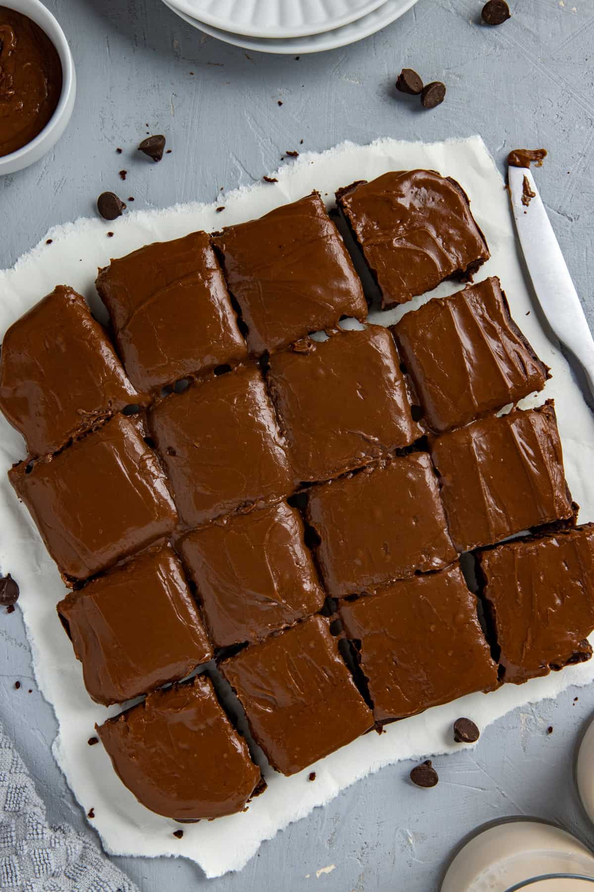 Baked and frosted brownies cut into 16 squares on white parchment paper with brownie crumbs scattered around.