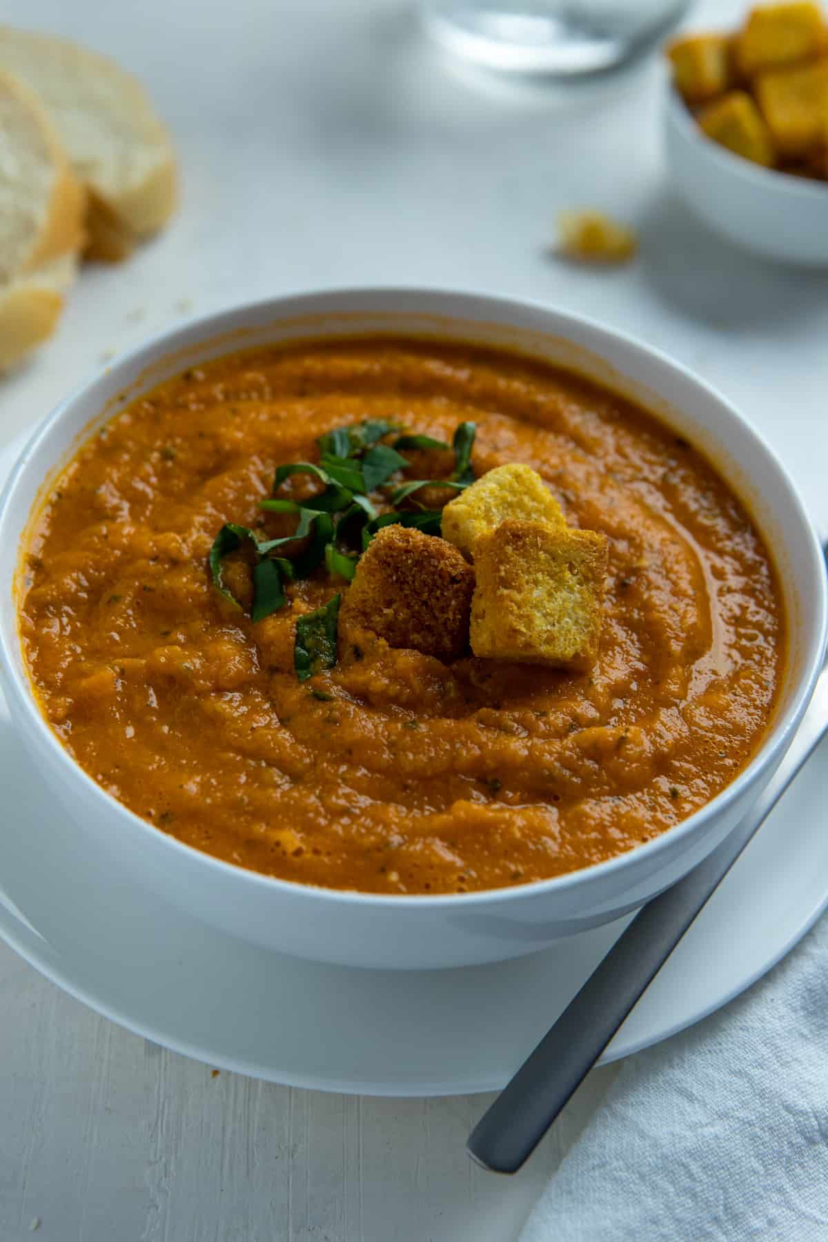 Tomato soup in white bowl with three croutons and fresh basil on top.