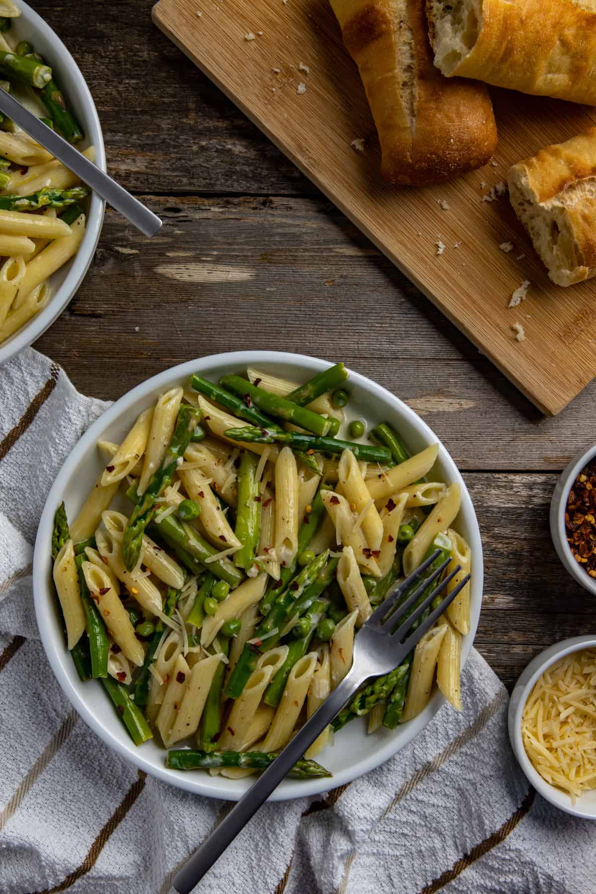 Cooked penne pasta with asparagus and sweet peas tossed in lemon garlic sauce and topped with parmesan cheese. In a white bowl with fork. French bread, extra cheese, and extra crushed red pepper in the background.