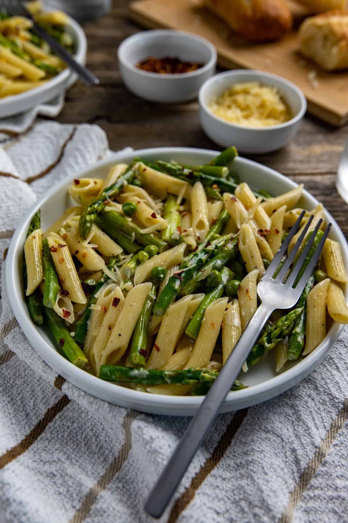 Cooked penne pasta with asparagus and sweet peas tossed in lemon garlic sauce and topped with parmesan cheese.  In a white bowl with fork.  French bread, extra cheese, and extra crushed red pepper in the background.