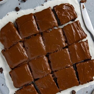 Baked and frosted brownies cut into 16 squares on white parchment paper with brownie crumbs scattered around.