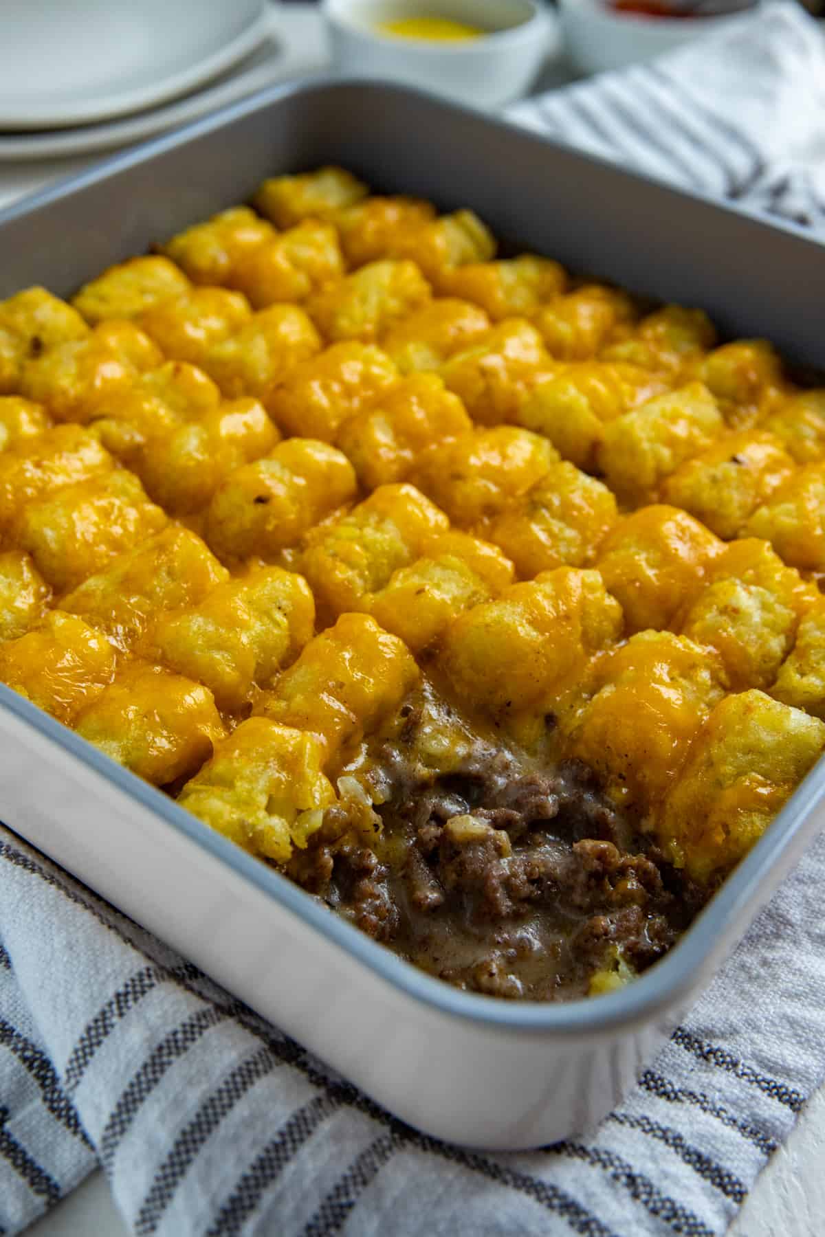 Baked tater tot casserole in square metal pan with scoop removed from corner. Mustard and ketchup in small round white bowls on the side.